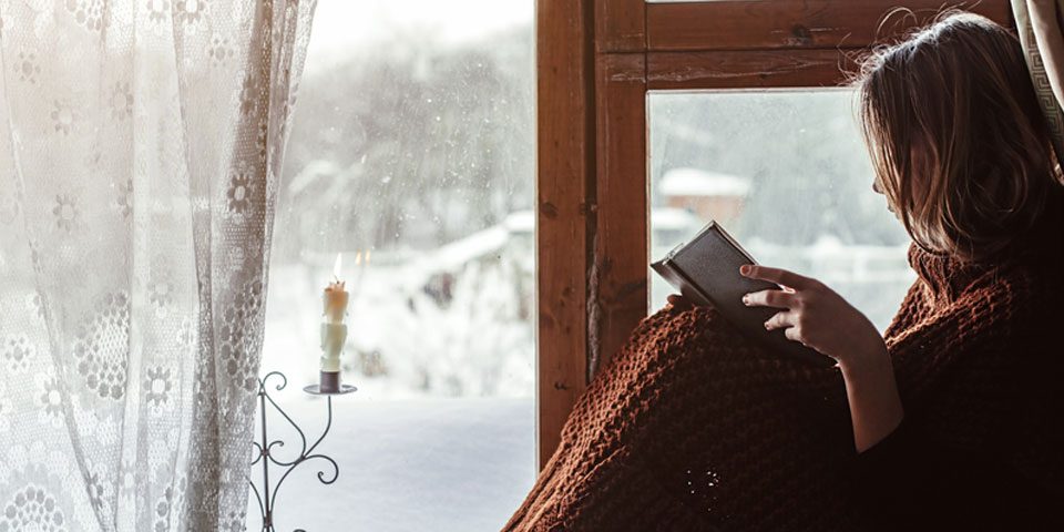 Lady Sat Next to Timber Windows Reading a Book, wearing a Blanket