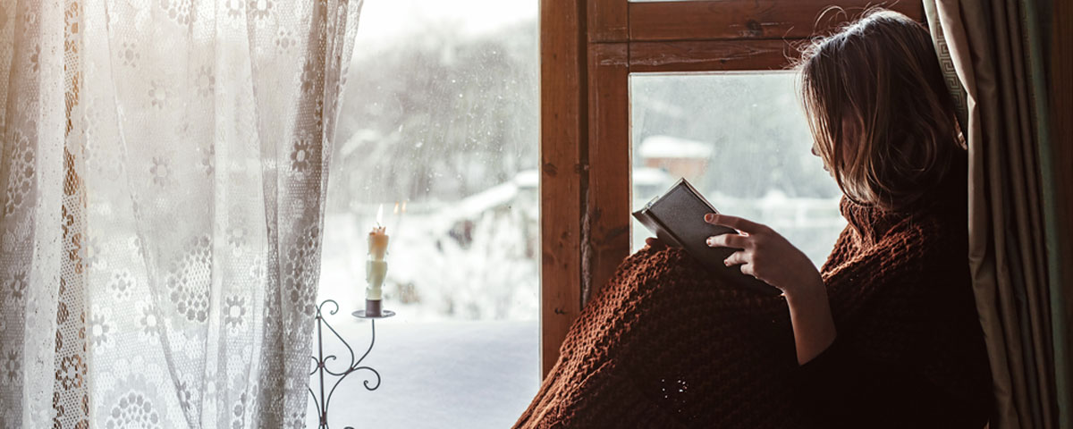 Lady Sat Next to Timber Windows Reading a Book, wearing a Blanket